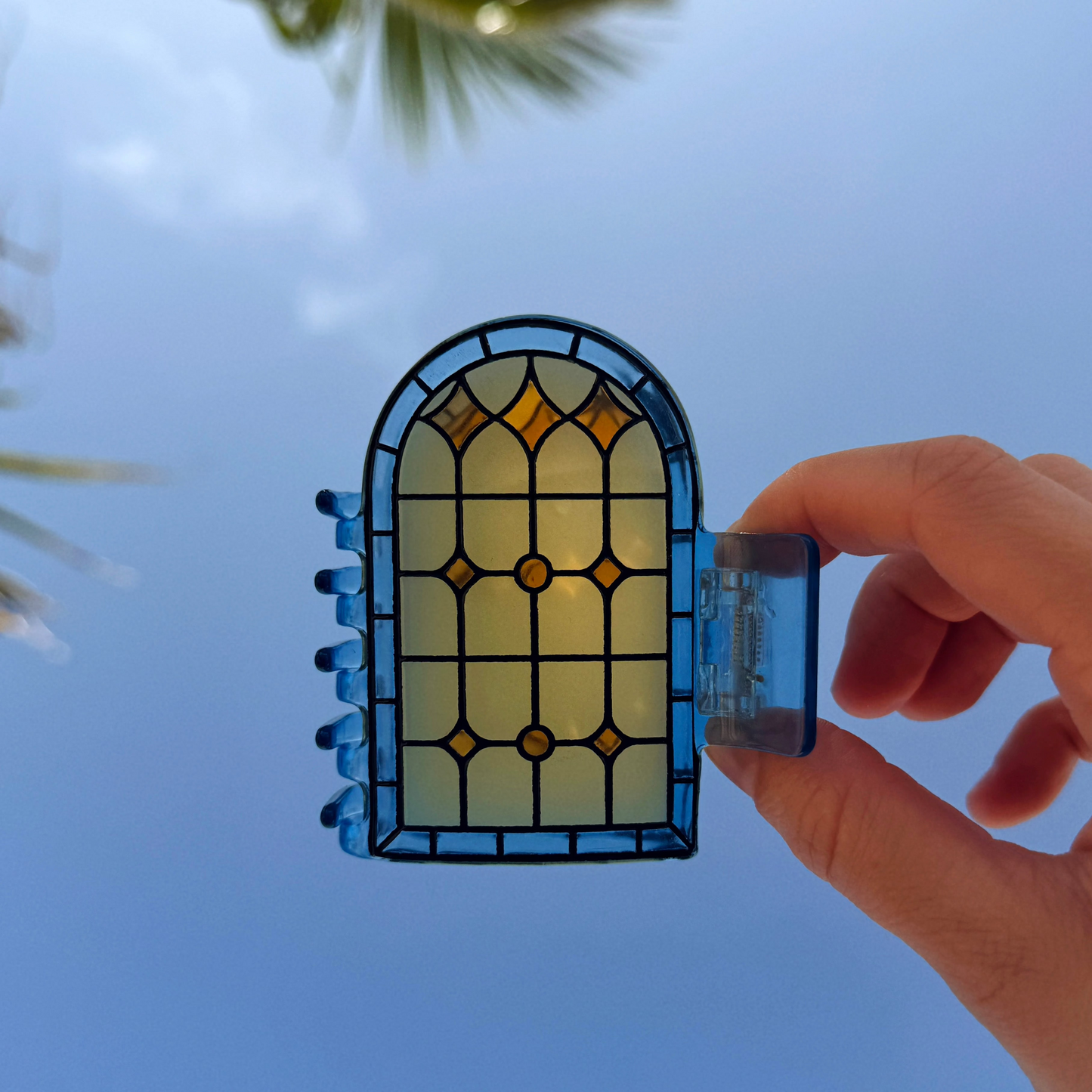 A hand holds a decorative stained-glass-style  claw clip shaped like an arched window panel against a clear blue sky with palm leaves in the background.