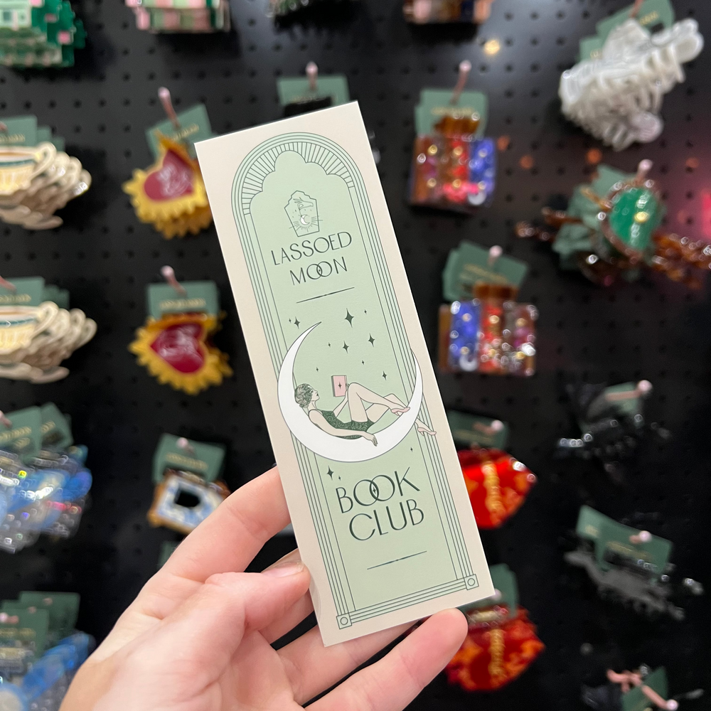 Hand holding a 'Lassoed Moon' Book Club bookmark with decorative background of small figurines.
