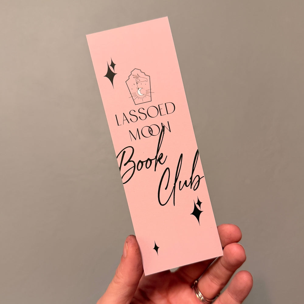 
                  
                    Pink bookmark with 'Lassoed Moon Book Club' text held by a hand against a gray background
                  
                