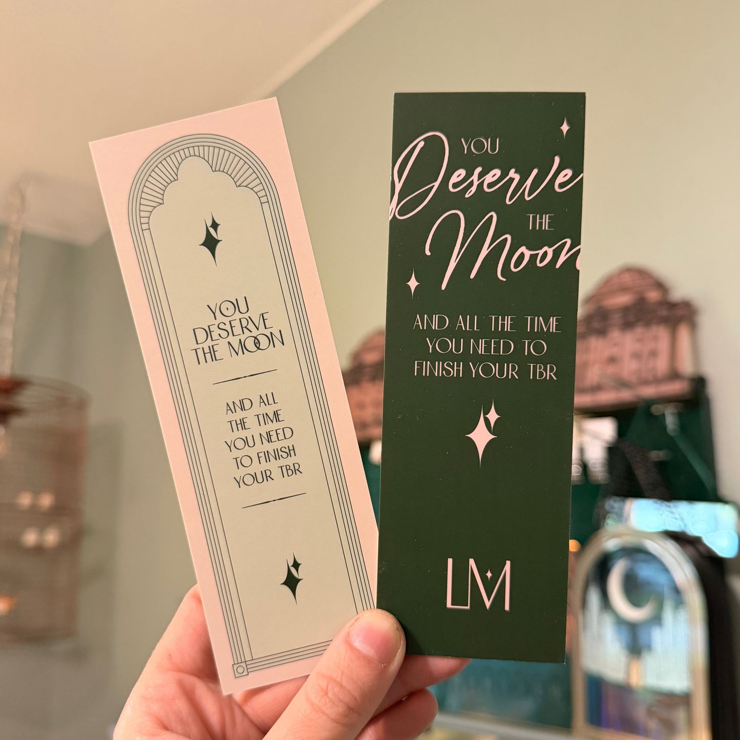Two bookmarks with motivational text held by a hand in front of a blurred indoor background.