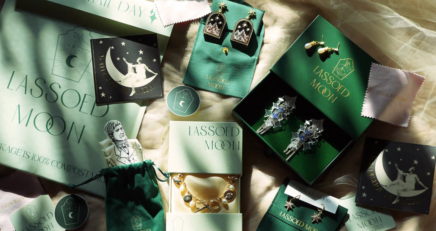 An above view of an arrangement of luxurious green jewelry packaging showing the logo Lassoed Moon with some jewelry pieces and hair accessories mixed throughout